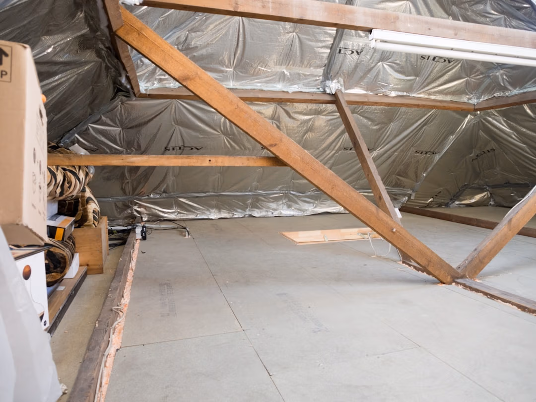 Attic space with exposed wooden trusses, silver insulation wrap, and stored boxes and supplies along the left wall